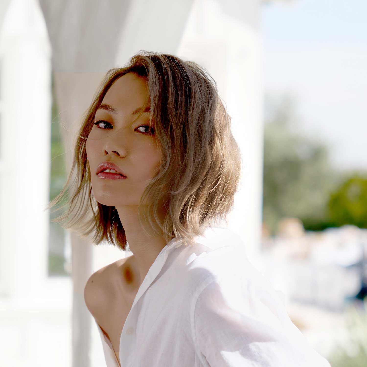 A woman with short, wavy blonde hair wearing a white shirt in an outdoor setting.