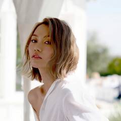 A woman with short, wavy blonde hair wearing a white shirt in an outdoor setting.