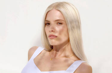 A person with long, straight blonde hair wearing a white top and a delicate gold necklace.