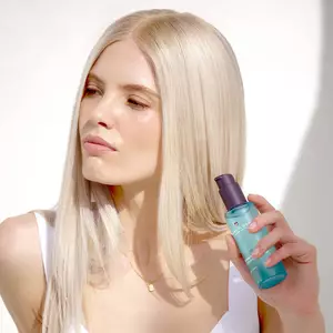 A person with long, straight blonde hair holding a bottle of Pureology Strength Cure Miracle Filler Treatment.