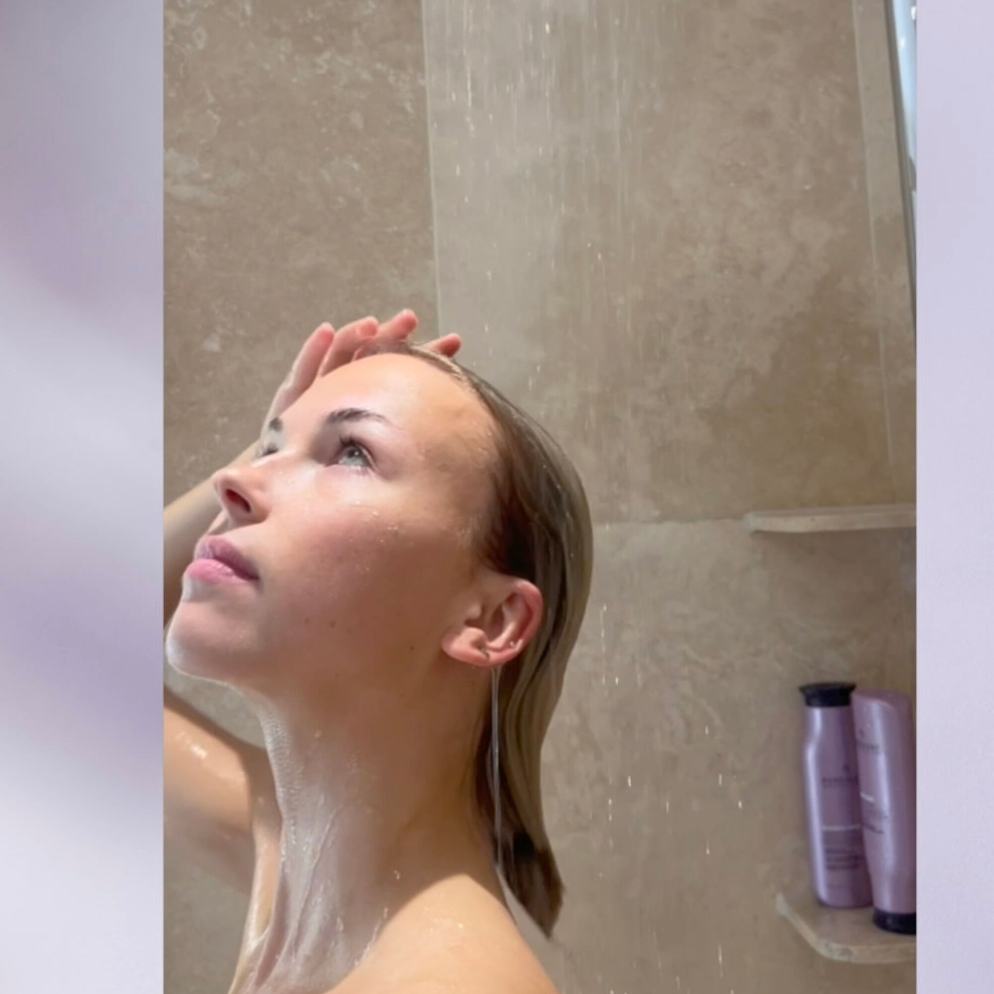 A person rinsing their hair in the shower with Pureology Hydrate bottles visible on a shelf.